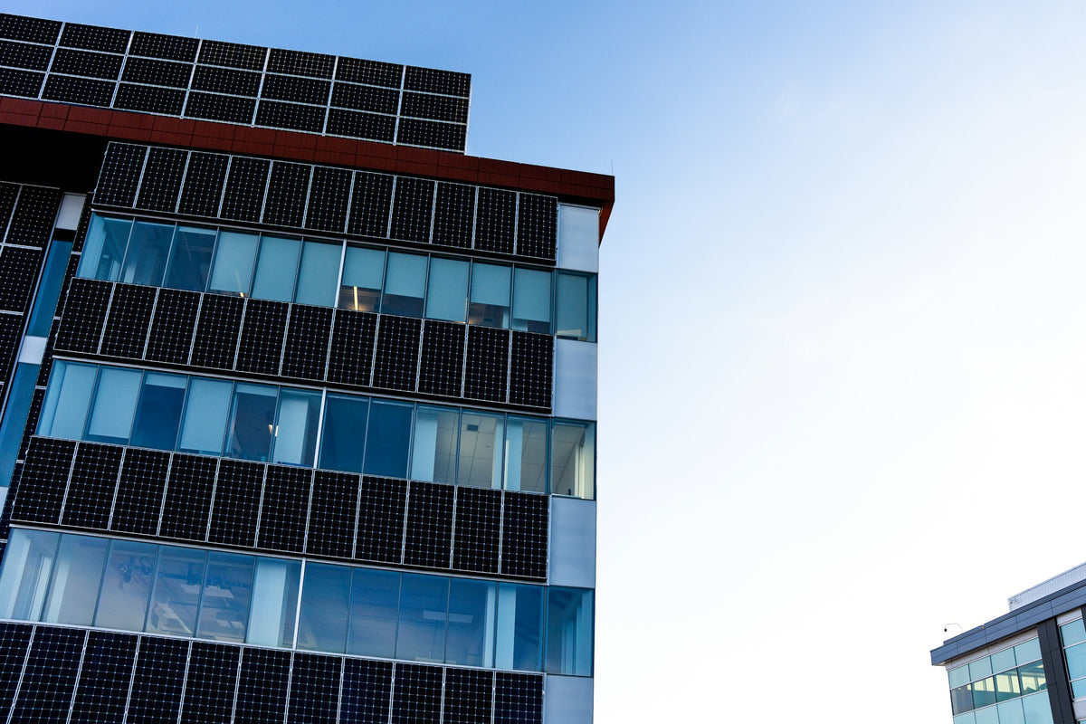 Modern building facade with rows of dark solar panels over blue-tinted glass windows, photographed against a clear blue sky.
