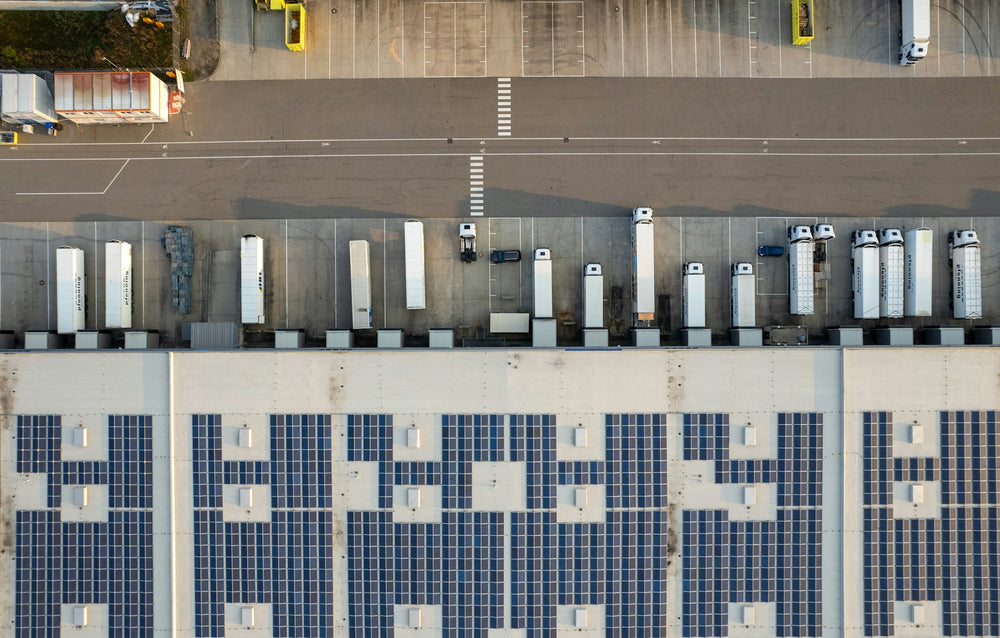 Aerial view of a large warehouse roof covered in rows of solar panels, with delivery trucks parked at the loading bays alongside.