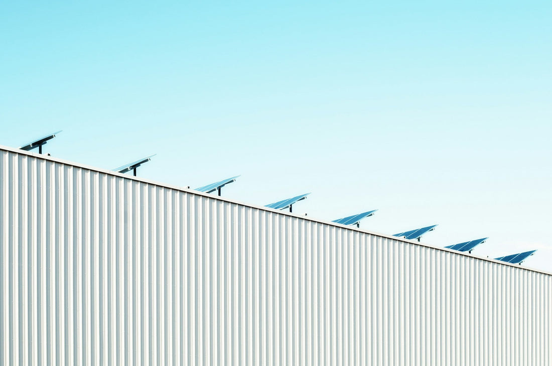 Row of tilted solar panels mounted along the roofline of a white corrugated-metal commercial building against a pale blue sky.
