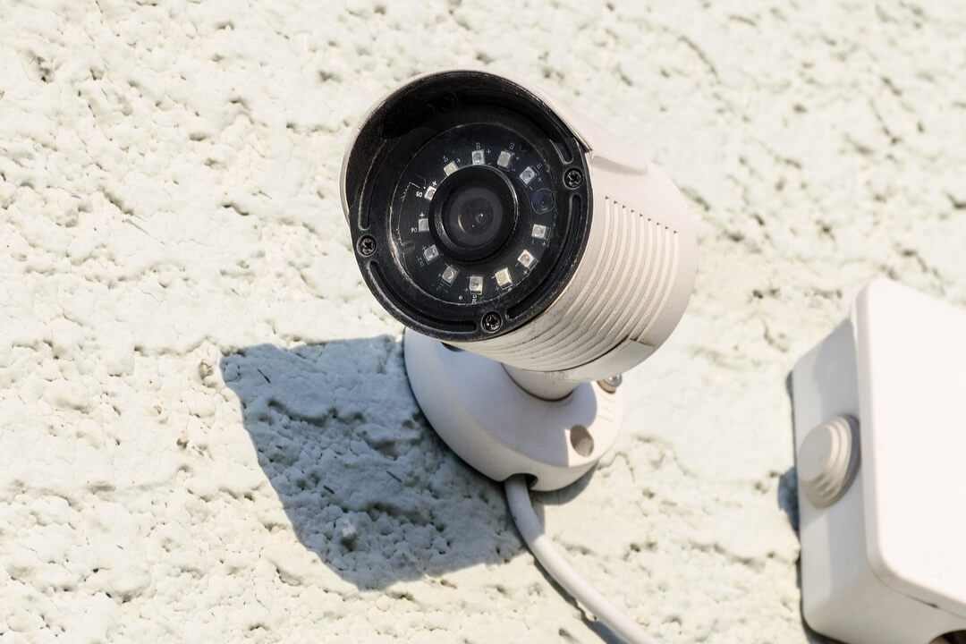 White CCTV security camera with infrared LEDs mounted on a textured white wall, casting a shadow in bright sunlight.