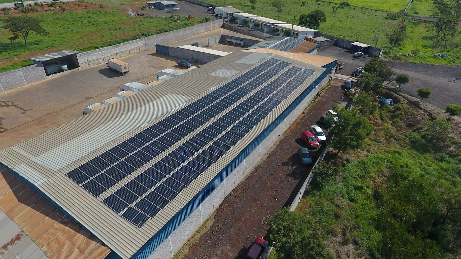 Aerial view of an industrial premises with commercial solar panels on its roof, surrounded by greenery and trucks.