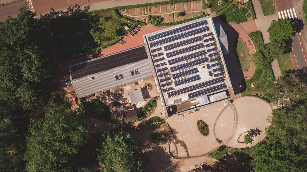 Aerial view of a large commercial solar panel installation covering the flat roof of a building, surrounded by trees and pathways.