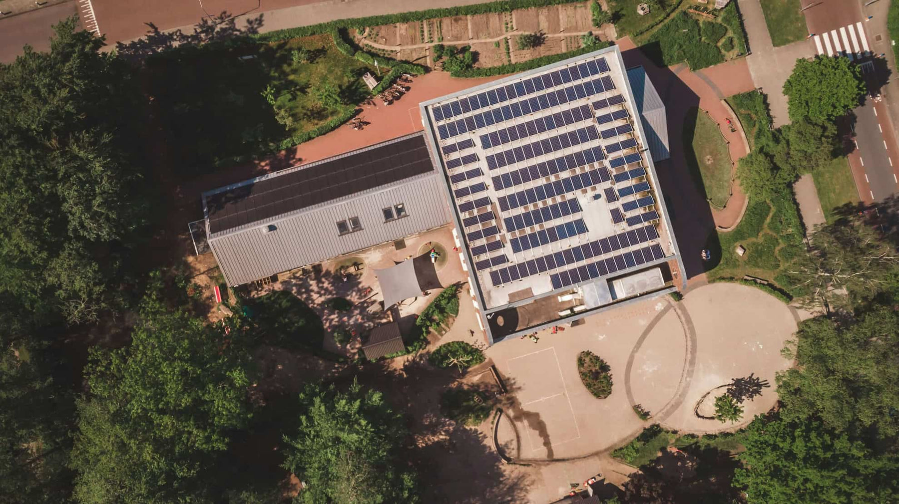Aerial view of a large commercial solar panel installation covering the flat roof of a building, surrounded by trees and pathways.