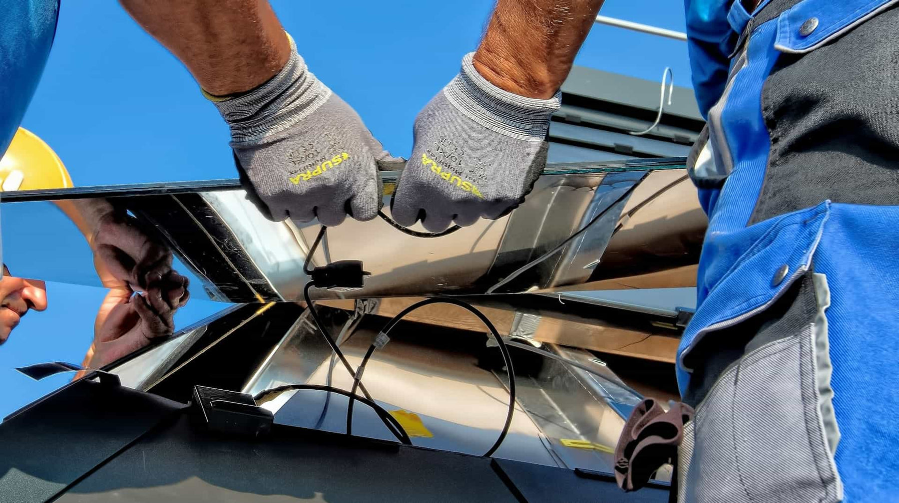 Person wearing gloves and a blue jacket working on a metal structure with a clear blue sky in the background.