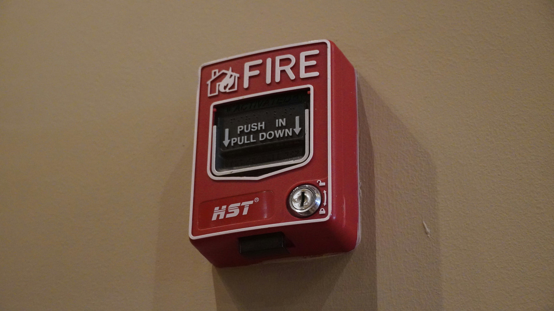 Red fire alarm push button on a beige wall with 'HST' branding.